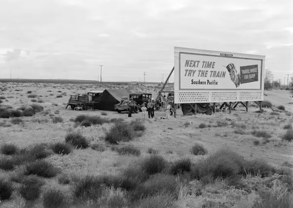 Photograph by Dorothea Lange during the Great Depression. Three families camped on the plains along the U.S. 99 in California. They are camped behind a billboard which serves as a partial windbreak. All are in need of work. 1938. The billboard says: ‘Next time try the train. Southern Pacific. Travel while you sleep’